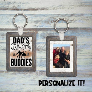 Puede incluir: Dos llaveros de madera con foto. Un marco tiene una foto de una familia de cuatro. El otro marco tiene el texto "Dad's Camping Buddies" con un diseño de montaña y fogata.