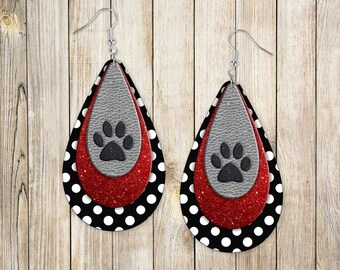 May include: A pair of dangle earrings with a layered design. The bottom layer is black with white polka dots, the middle layer is red glitter, and the top layer is gray with a black paw print.