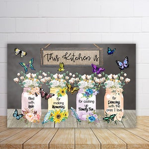 May include: A kitchen wall art print with a gray background and a sign that reads "This Kitchen is". Four mason jars with floral designs and butterflies are arranged in a row. Each jar has a different saying: "filled with love", "for making memories", "for cooking up family fun", and "for dancing with the ones I love".