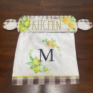 Puede incluir: Una toalla de cocina blanca con un borde a cuadros marrón y blanco. La toalla tiene un diseño de corona de limón con una gran letra "M" en el centro. La toalla está colgada de un rodillo de madera con la palabra "KITCHEN" escrita en él.