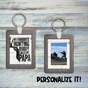 Puede incluir: Dos llaveros con marco de madera y fondo blanco. El llavero de la izquierda tiene una silueta en blanco y negro de una cabeza de ciervo con el texto "My Favorite Hunting Buddy Calls Me Papa". El llavero de la derecha tiene una foto de dos personas cazando en un campo.