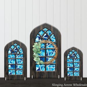 May include: Three wooden stained glass window frames with a blue and white stained glass design. The largest frame has a eucalyptus wreath hanging on it.