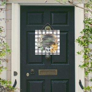 Könnte beinhalten: Ein schwarz-weiß karierter Türkranz mit einem Kranz aus Herbstblättern und Kürbissen. Der Kranz befindet sich auf einem rustikalen Holzgrund.