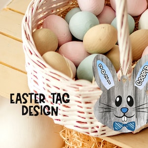 Könnte beinhalten: Ein grauer Osteranhänger in Hasenform mit blauen Ohren und einer blauen Schleife. Der Anhänger hat den Text "COOPER" auf einem Ohr und "2021" auf dem anderen Ohr. Der Anhänger befindet sich vor einem Korb mit pastellfarbenen Ostereiern.