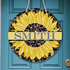 May include: A round door hanger with a black and gold glitter background featuring a yellow sunflower with a gray banner that reads "SMITH".