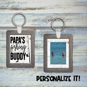 Puede incluir: Dos llaveros de fotos de madera con acabado blanqueado. Un llavero presenta el texto "Papa's fishing buddy" con un gráfico de caña de pescar. El otro llavero tiene una foto de dos personas en el agua. Ambos llaveros tienen un anillo de metal plateado.