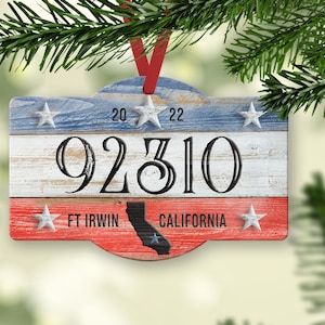 Op de afbeelding: Een houten kerstversiering met een rood, wit en blauw Amerikaans vlagontwerp. De versiering heeft de tekst "92310 FT IRWIN CALIFORNIA" en een silhouet van de staat Californië.