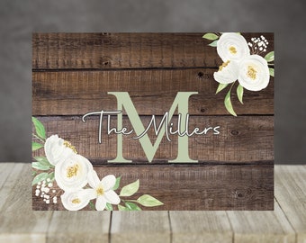May include: A wooden sign with a rustic brown background and white flowers. The sign says "The Millers" with a large green "M" in the center.
