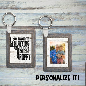 Puede incluir: Dos llaveros con marco de madera. Un llavero presenta un fondo blanco con una silueta de ciervo negro y el texto "My Favorite Hunting Buddy Calls Me Poppy". El otro llavero presenta una foto de dos personas sonriendo.