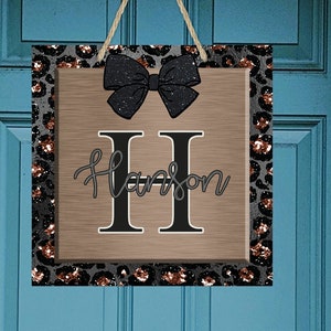May include: A wooden door hanger with a black and gold leopard print border. The center features a large black bow and the letter "H" with the name "Harrison" written below.