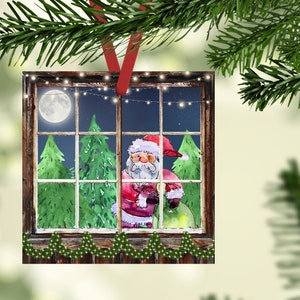 May include: A Christmas ornament featuring a watercolor illustration of Santa Claus peeking through a window with a bag of gifts. The window is framed in wood and has a string of lights hanging above it. The background is a dark blue night sky with a full moon and stars.