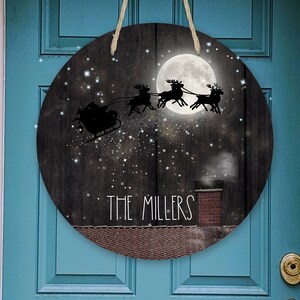 May include: A round wooden door hanger with a black and white illustration of Santa Claus flying in a sleigh pulled by reindeer. The moon is full and there are stars in the sky. The text "THE MILLERS" is written on the bottom of the hanger.