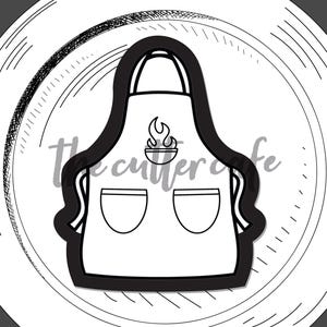 Può includere: Tagliabiscotti a forma di grembiule in bianco e nero. Il design del grembiule include due tasche e una grafica a fiamma sopra le tasche. Il testo "apron" è in basso. Il logo The Cutter Cafe è in alto a destra.
