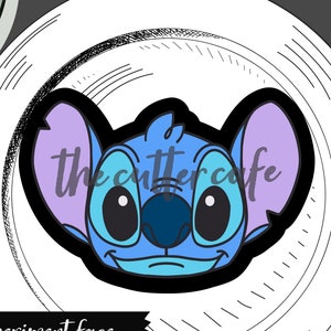 May include: Blue cookie cutter in the shape of Stitch's face from the Disney movie Lilo & Stitch. The cutter is on a white plate with a black and white background. The text "the cutter cafe" is visible in the image.