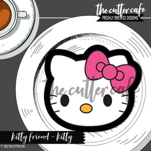 May include: A black and white cookie cutter in the shape of a cartoon cat face with a pink bow. The cutter is on a white plate with a coffee cup in the upper left corner. Text on the image reads "the cutter cafe" and "kitty friend - kitty".