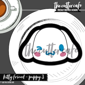 May include: A black and white cookie cutter shaped like a puppy's head with blue and pink details. The cutter is on a white plate with the text "the cutter cafe" in black and white.