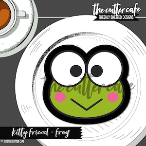 Może przedstawiać: Wycinarka do ciasteczek w kształcie żaby w kolorze czarnym i zielonym z dużymi oczami i różowymi policzkami. Wycinarka znajduje się na białym talerzu z czarno-białą wzorzystą obwódką. Pod talerzem znajduje się tekst "kitty friend - frog".