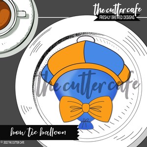May include: A blue and orange bow tie balloon cookie cutter. The cutter is shaped like a balloon with a bow tie. The text "the cutter cafe" is visible in the background.