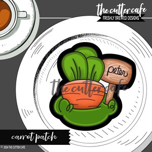 May include: A green carrot cookie cutter with a brown sign that says "Peter" and green leafy tops. The cookie cutter is on a white plate with a black and white patterned border.
