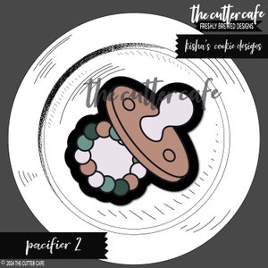 May include: A cookie cutter in the shape of a pacifier with a beaded necklace attached. The pacifier is brown and white, and the beads are green, white, and brown.