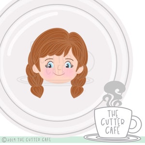 Puede incluir: Un plato de cerámica blanco con la cara de una niña de dibujos animados con cabello castaño y ojos azules. La niña sonríe y tiene un rubor rosado en las mejillas. El plato está sobre una superficie verde con una taza de café blanca con el texto "THE CUTTER CAFE" en negro.