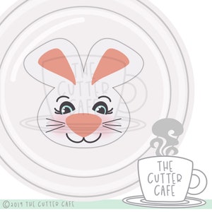May include: A white bunny face with pink ears and nose, and blue eyes. The bunny is looking at a steaming cup of coffee with the text "THE CUTTER CAFE" on it.