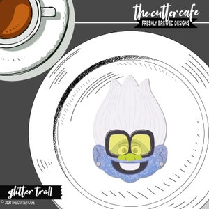 Può includere: Un tagliapasta a forma di troll blu glitterato con capelli bianchi e occhiali gialli. Il tagliapasta è su un piatto bianco con un design bianco e nero. Il testo "the cutter cafe" e "freshly brewed designs" è nella parte superiore dell'immagine. Il testo "glitter troll" e "© 2020 THE CUTTER CAFE" è nella parte inferiore dell'immagine.