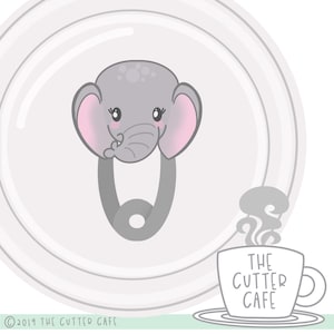Peut inclure: Un motif de sucette pour bébé en forme d'éléphant gris avec des oreilles roses et un visage souriant. La sucette est conçue comme une épingle de sûreté. L'image comprend une tasse à café avec le texte "THE CUTTER CAFE" et le copyright "© 2019 THE CUTTER CAFE".