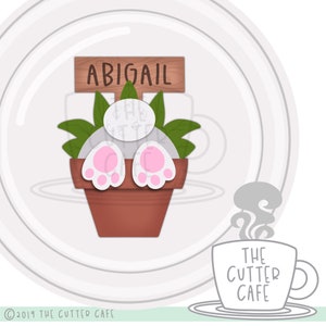 May include: A white bunny rabbit with pink feet is sitting in a brown flower pot with green leaves. The pot has a wooden sign that says "ABIGAIL" on it. The image is set against a white background with a gray circle around the bunny.