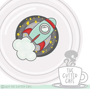 Może przedstawiać: Biały ceramiczny kubek z napisem "THE CUTTER CAFE" czarnymi literami. Kubek stoi na zielonej powierzchni z białym talerzem i kreskówkowym statkiem kosmicznym w kosmicznej scenerii.