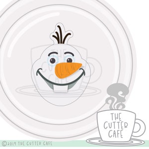 May include: A white cookie cutter in the shape of Olaf's face from the movie Frozen. The cutter has a black hair, brown eyes, a carrot nose, and a big smile. The cutter is on a white plate with a gray mug with the text "THE CUTTER CAFE" on it.