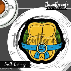 May include: A yellow and green turtle shell cookie cutter with a blue ribbon and the number 5 on it. The cookie cutter is on a white plate with a black and white background. The text "turtle tummy" is below the plate. The text "the cutter cafe" is in the upper right corner of the image.
