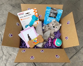 May include: A brown cardboard box filled with dog toys and treats. The box is decorated with purple paw prints. The toys include a blue bone, a grey squirrel plush toy, a cork bone, a purple and white tube, and a green tennis ball. The treats are in a blue bag with the text "American Journey Training Bites Soft".