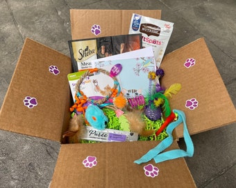 Puede incluir: Una caja de cartón marrón llena de juguetes para gatos, golosinas y una tarjeta de felicitación. La caja está decorada con pegatinas moradas de huellas de patas. Contenido: juguete de ratón azul, juguetes de plumas y paquetes de comida para gatos.