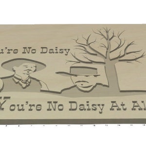 Peut inclure: Plaque en bois avec une &oelig;uvre d'art grav&eacute;e sur le th&egrave;me de l'Ouest. Le dessin repr&eacute;sente deux personnages avec des chapeaux et un arbre, avec le texte "You're No Daisy" et "You're No Daisy At All" grav&eacute; dans le bois. La plaque est marron clair.