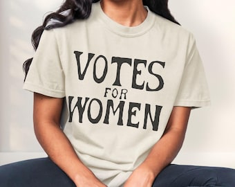 Puede incluir: Una camiseta color crema con las palabras "VOTES FOR WOMEN" impresas en letras negras y en negrita. La camiseta tiene mangas cortas y cuello redondo. La persona lleva pantalones azul oscuro.