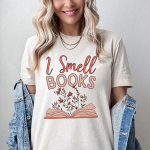 May include: A beige t-shirt with the text "I Smell Books" in pink and a graphic of an open book with flowers growing from it.