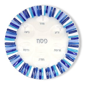 May include: A round glass Seder plate featuring a border of small, rectangular blue, light blue, and white glass tiles. The center of the clear plate displays six circular indentations, each labeled with Hebrew text, and the word Pesach in the center.