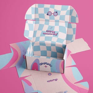 May include: A cardboard shipping box with a light blue and white checkerboard pattern on the interior. The box features pink, blue, and purple wavy designs on the exterior. Text on the box reads 7,000 tiny reasons to bling! and Blingkitybites.