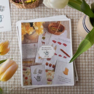 Peut inclure: Une pile de cartes illustrées avec le texte "Morning Moments" et "spring" sur une surface à carreaux. Les cartes présentent des illustrations à l'aquarelle d'outils de jardinage et de fleurs. Des tulipes jaunes et un panier de crayons sont également visibles.