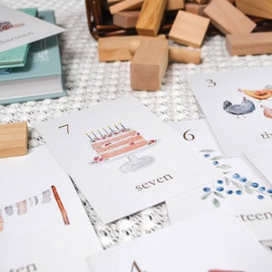 Puede incluir: Un juego de cartas blancas con ilustraciones y n&uacute;meros. Las cartas presentan im&aacute;genes de un pastel de cumplea&ntilde;os, pollos, ar&aacute;ndanos, ropa en un tendedero y un plato de comida. Los n&uacute;meros en las cartas son 3, 5, 6, 7, 10 y 13. Las palabras "tres", "siete" y "trece" tambi&eacute;n son visibles en las cartas.