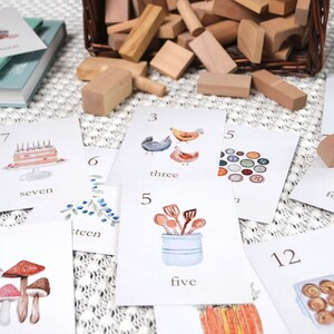 Puede incluir: Un conjunto de tarjetas de memoria coloridas con n&uacute;meros e ilustraciones correspondientes. Los n&uacute;meros van del 3 al 18. Las ilustraciones incluyen un pastel de cumplea&ntilde;os, pollos, un cubo de cucharas, champi&ntilde;ones, una corona de bayas, un rastrillo, un plato de pasteles y una pila de palos. Las tarjetas est&aacute;n sobre una superficie blanca con una cesta de mimbre llena de bloques de madera en el fondo.