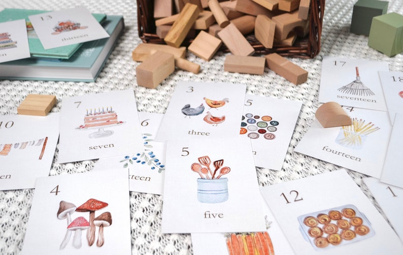 Puede incluir: Un conjunto de tarjetas de memoria coloridas con n&uacute;meros e ilustraciones correspondientes. Los n&uacute;meros van del 3 al 18. Las ilustraciones incluyen un pastel de cumplea&ntilde;os, pollos, un cubo de cucharas, champi&ntilde;ones, una corona de bayas, un rastrillo, un plato de pasteles y una pila de palos. Las tarjetas est&aacute;n sobre una superficie blanca con una cesta de mimbre llena de bloques de madera en el fondo.