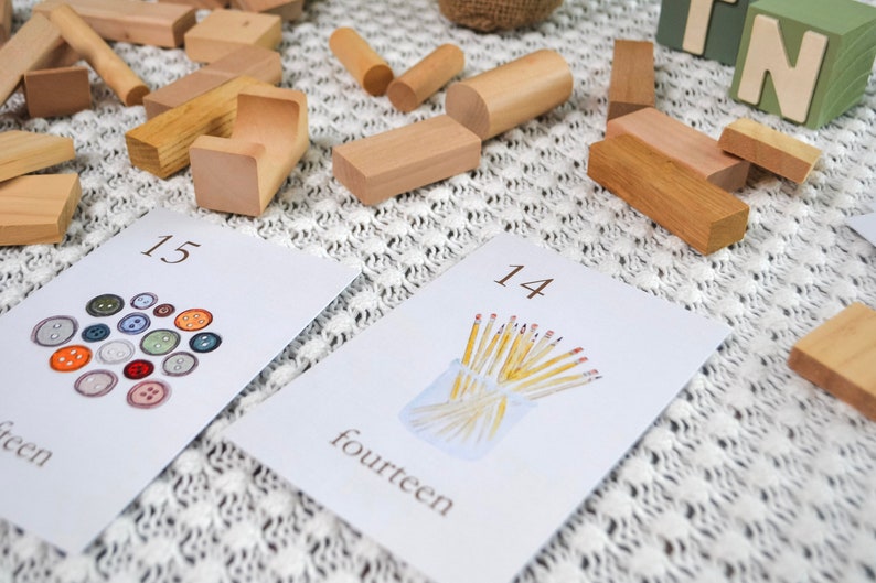 Puede incluir: Dos tarjetas blancas con n&uacute;meros y palabras en ellas. La tarjeta de la izquierda muestra el n&uacute;mero 15 y la palabra "quince" y tiene una imagen de 15 botones de diferentes colores. La tarjeta de la derecha muestra el n&uacute;mero 14 y la palabra "catorce" y tiene una imagen de 14 l&aacute;pices amarillos en un frasco de vidrio.