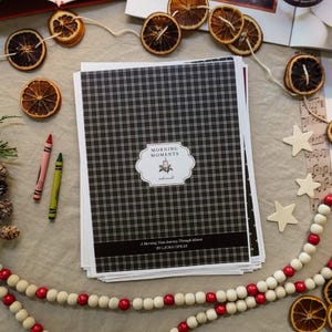 Puede incluir: Un conjunto de cuadernos con un patrón de cuadros blancos y negros y el texto "MORNING MOMENTS". La imagen también incluye rodajas de naranja secas, crayones y cuentas decorativas, creando un ambiente festivo.