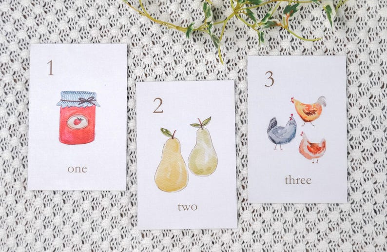 Puede incluir: Tres tarjetas con ilustraciones de acuarela y n&uacute;meros. La primera tarjeta muestra un tarro de mermelada con el n&uacute;mero 1 y la palabra "uno". La segunda tarjeta muestra dos peras con el n&uacute;mero 2 y la palabra "dos". La tercera tarjeta muestra tres pollos con el n&uacute;mero 3 y la palabra "tres".