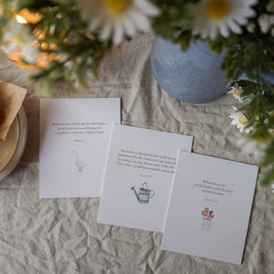 May include: Three white cards with illustrations and text, alongside a partially eaten muffin on a plate. The cards feature a goose, watering can, and flowers. Daisies and a blue vase are in the background.