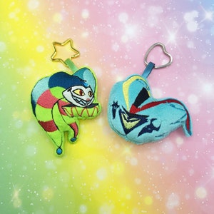 May include: Two plush keychains, one green and one blue, with embroidered details. The green keychain is shaped like a jester with a red and white striped hat and a mischievous grin. The blue keychain is shaped like a heart with a star and a lightning bolt design.