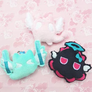 Op de afbeelding: Drie pluche speelgoedfiguren worden getoond. Eén is lichtroze met witte vleugels, een andere is lichtblauw met witte vleugels en de derde is zwart met roze en turquoise accenten. Ze liggen op een roze bloemenachtergrond.