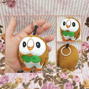 May include: A brown and white plush keychain of a cartoon owl with green leaves around its neck. The owl has large, round eyes and a small, orange beak. The keychain is attached to a silver ring.